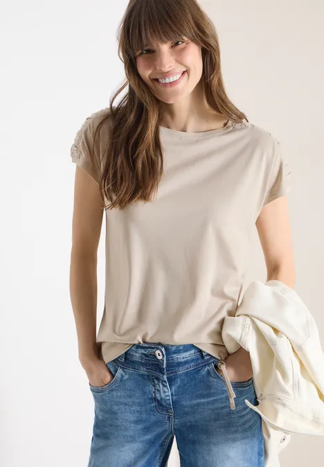 Eine Person steht in blauen Jeans und einem beigen T-Shirt, die Hände in den Taschen, mit einer leichten Jacke über dem Arm.